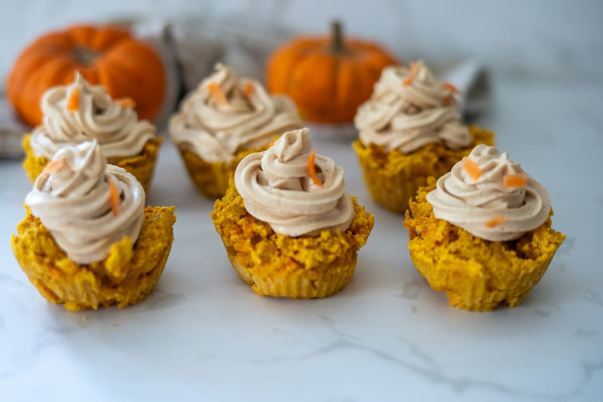 Seven pumpkin pupcakes with light brown frosting are arranged on a white surface, with two small pumpkins blurred in the background.