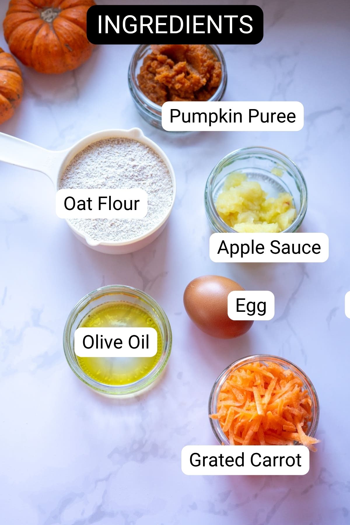 Labeled ingredients for pumpkin pupcakes on a white surface: oat flour, pumpkin puree, applesauce, egg, olive oil, and grated carrot, with small pumpkins in the background.