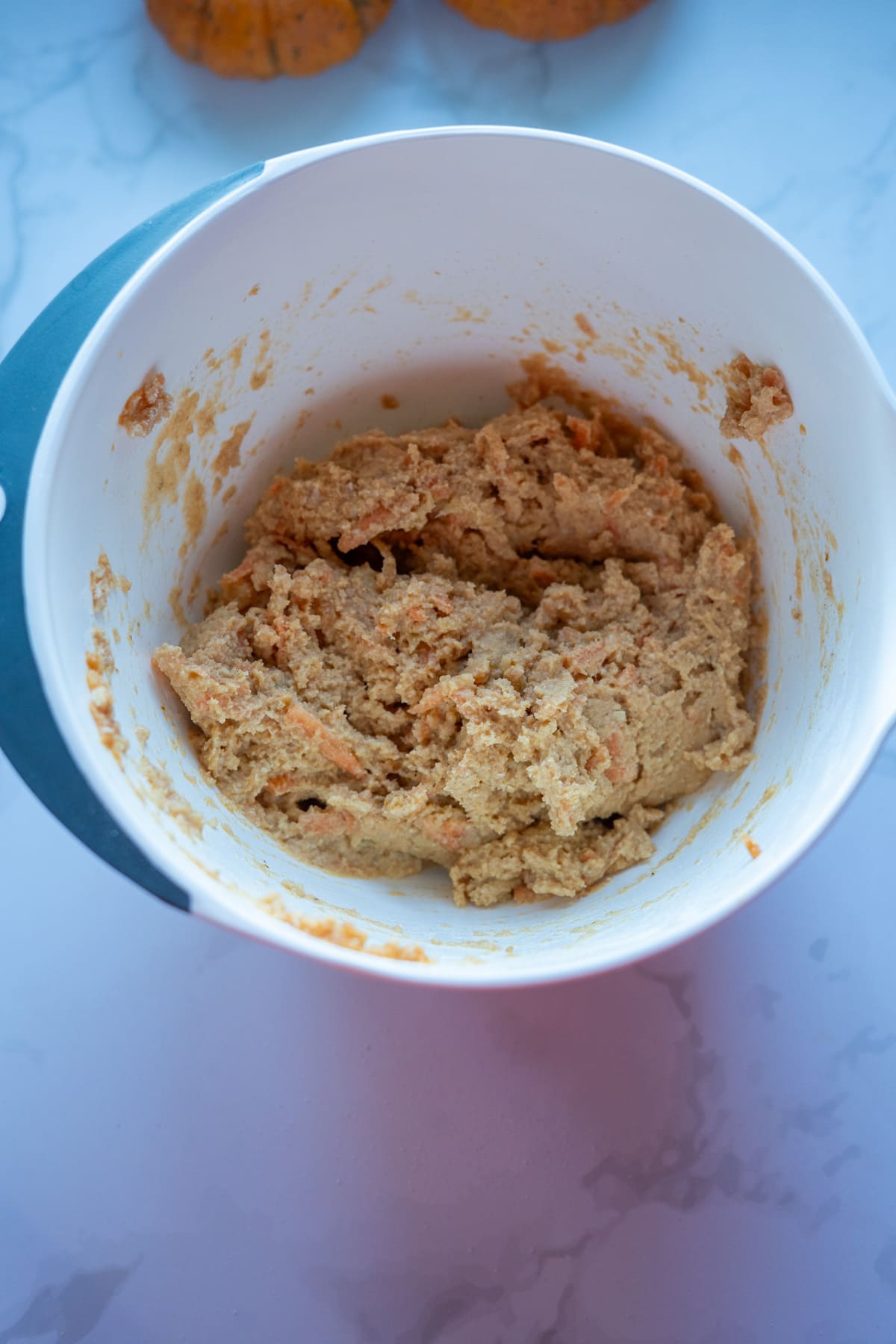 A white mixing bowl containing a thick, beige pumpkin pupcakes dough mixture sits on a marble countertop.
