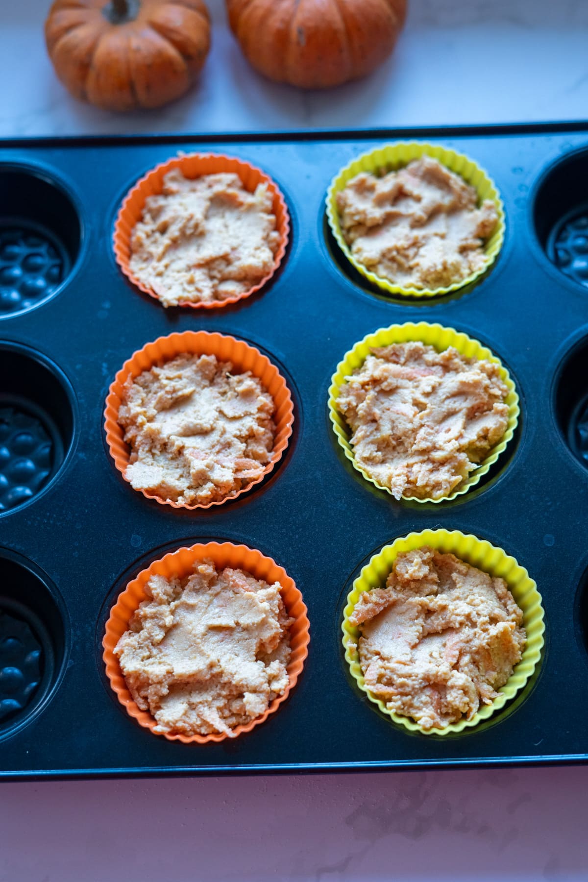 A muffin tin with six silicone baking cups filled with unbaked pumpkin pupcakes batter sits on a counter, with two small pumpkins in the background.