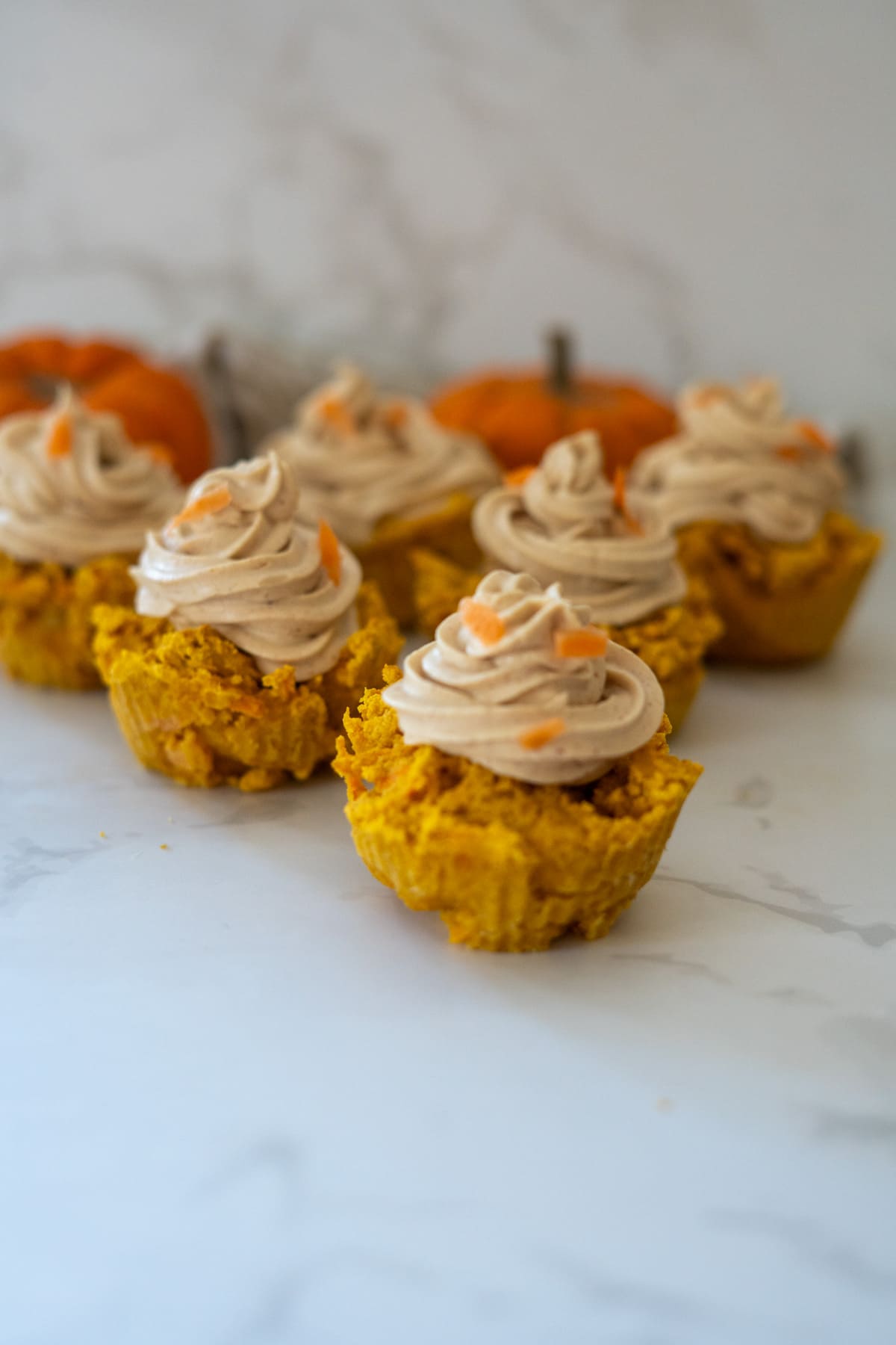 Six pumpkin muffins, known as pumpkin pupcakes, topped with swirled frosting are arranged on a white surface with small pumpkins in the blurred background.