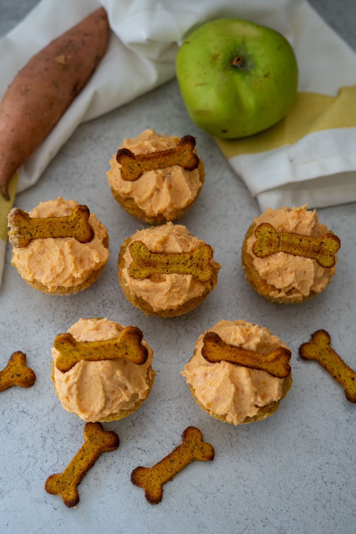 Six dog cupcakes with light orange frosting and bone-shaped dog treats on top, arranged on a surface with a sweet potato, green apple, and a cloth in the background.