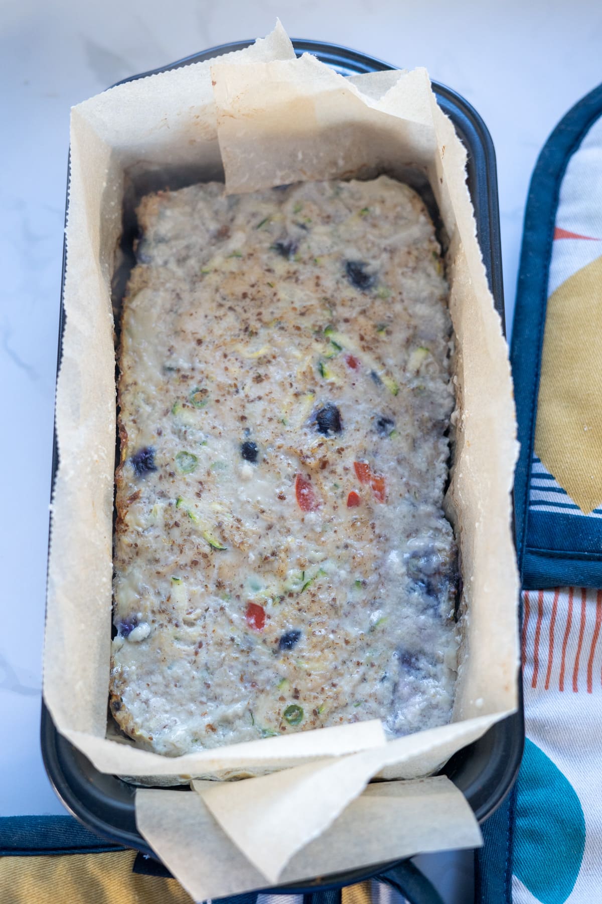 A loaf pan lined with parchment paper holds unbaked chicken meatloaf for dogs, featuring visible blueberries and vegetable pieces, beside a colorful oven mitt.
