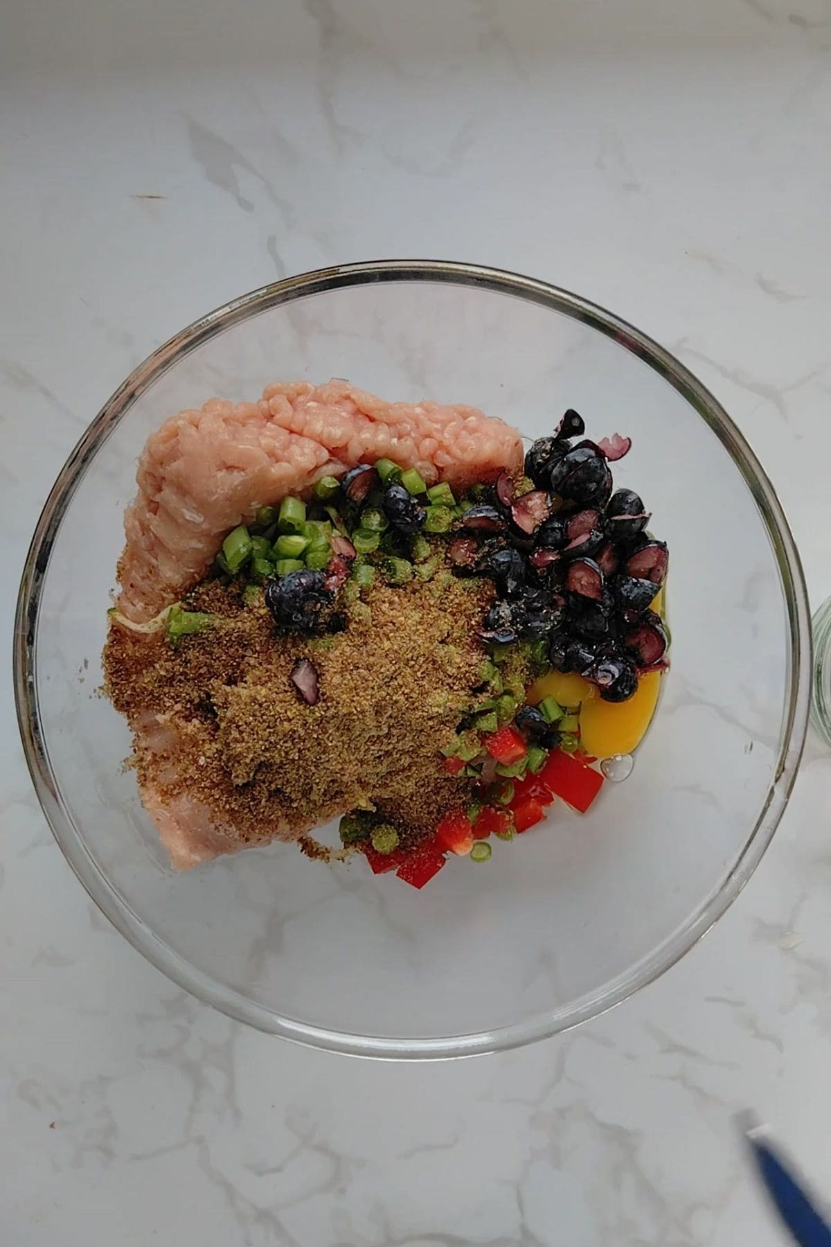A glass bowl containing ground meat for chicken meatloaf for dogs, an egg, chopped green and red vegetables, blueberries, and brown seasoning on a white marble surface.