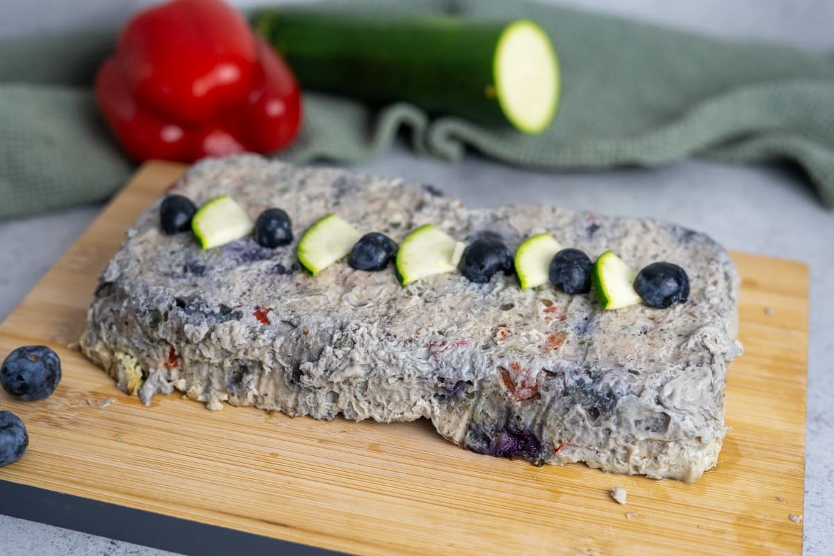 A rectangular loaf with a grayish texture and blueberries and zucchini slices on top, placed on a wooden cutting board with a red bell pepper and zucchini in the background.