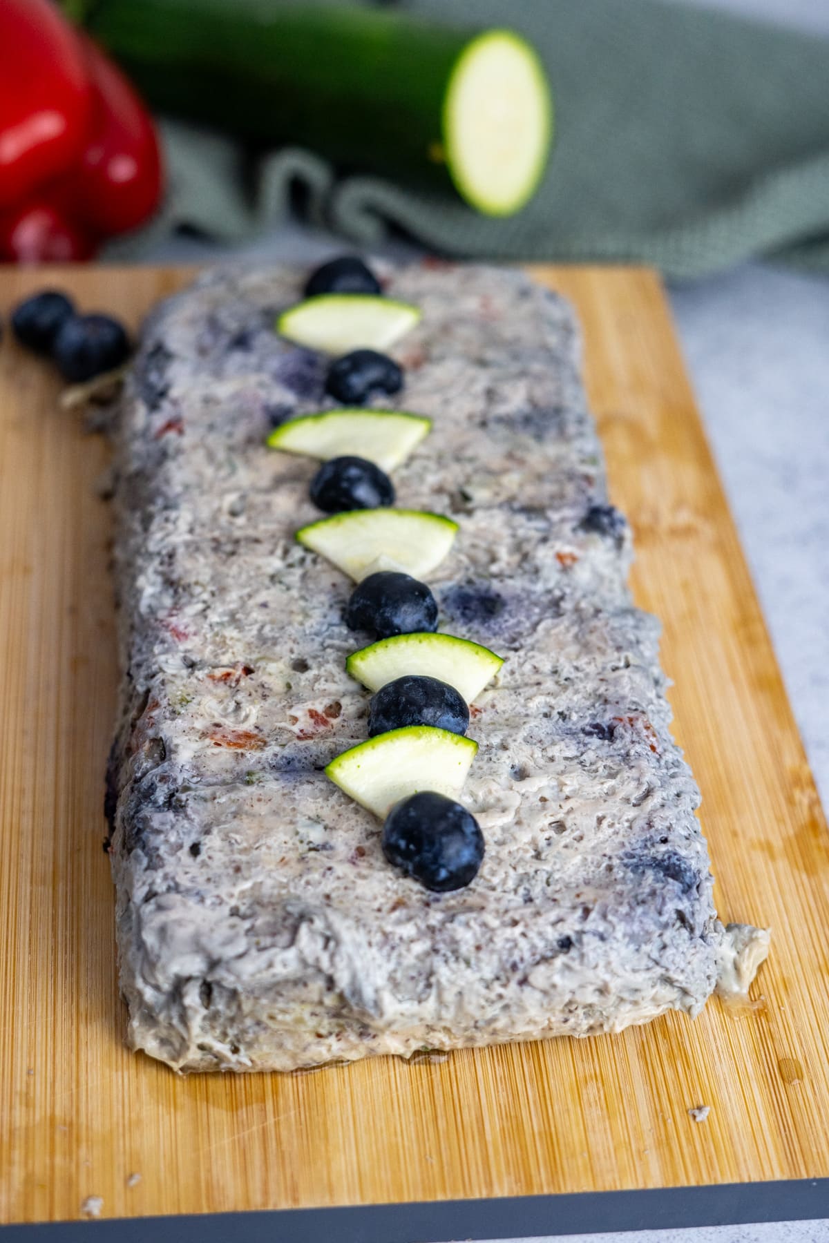 A rectangular chicken meatloaf for dogs with a grayish, textured surface is garnished with alternating slices of zucchini and whole blueberries, displayed on a wooden cutting board.