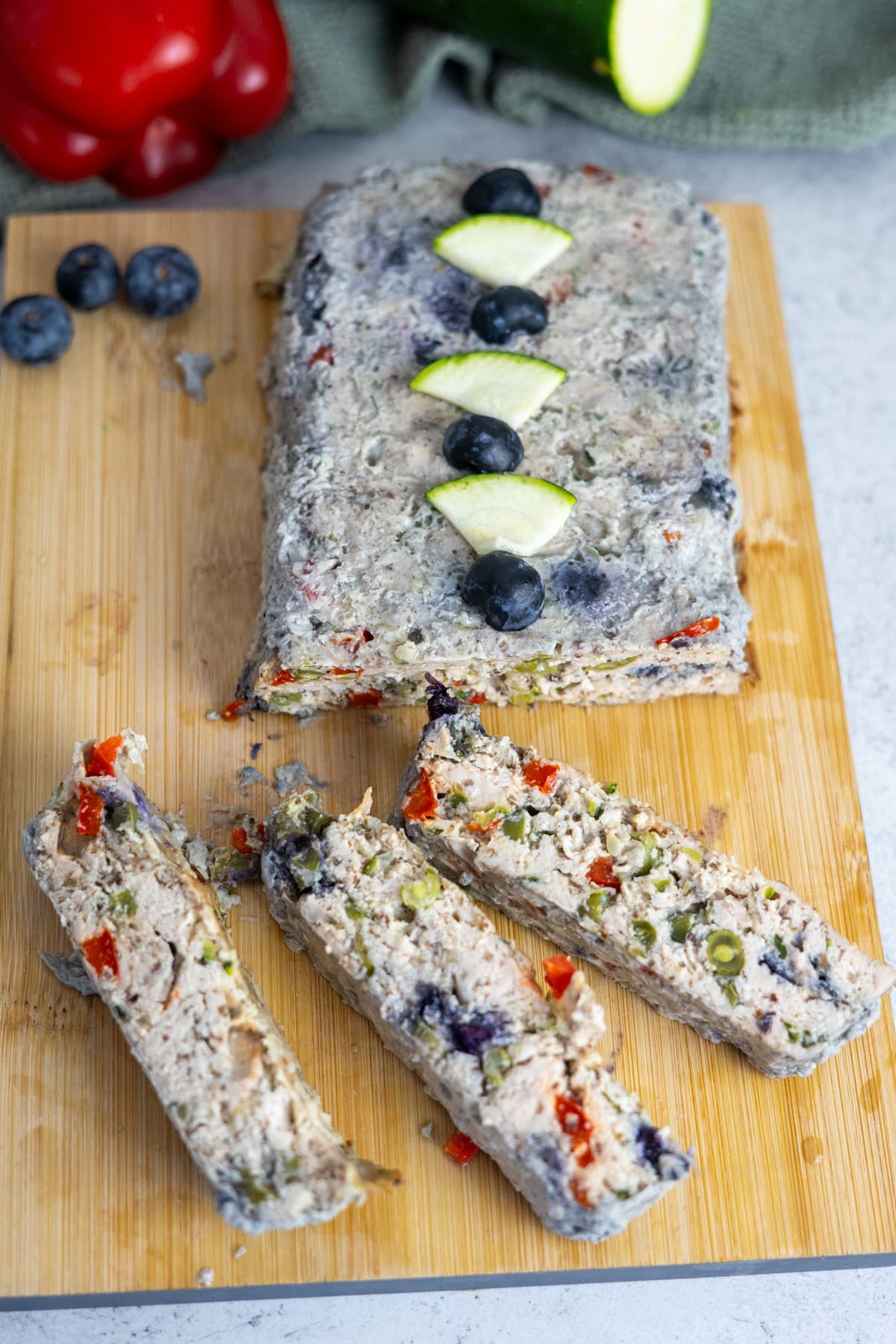 A loaf of chicken meatloaf for dogs with visible pieces of vegetables and blueberries is sliced on a wooden board, garnished with blueberry and cucumber slices. Red bell pepper and zucchini are in the background.