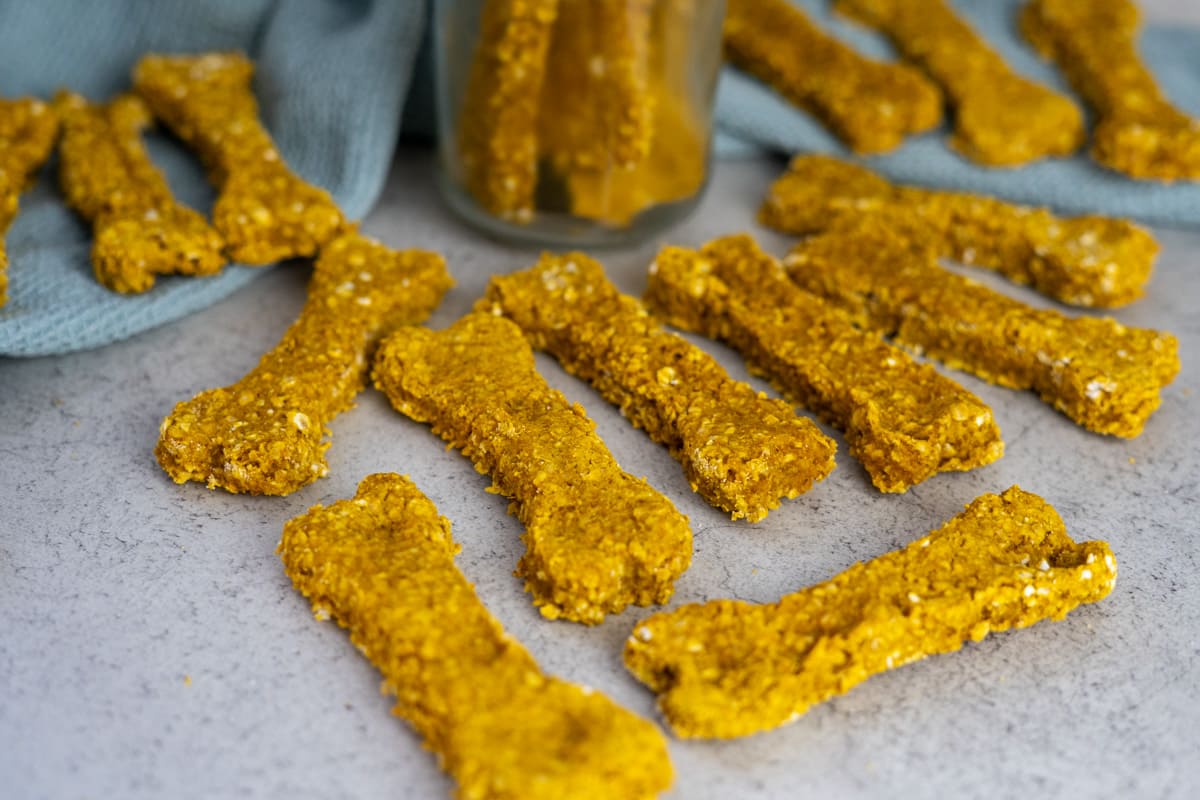 Several bone-shaped, yellow-orange dog treats are arranged on a light surface, with some leaning against a jar and others scattered around.