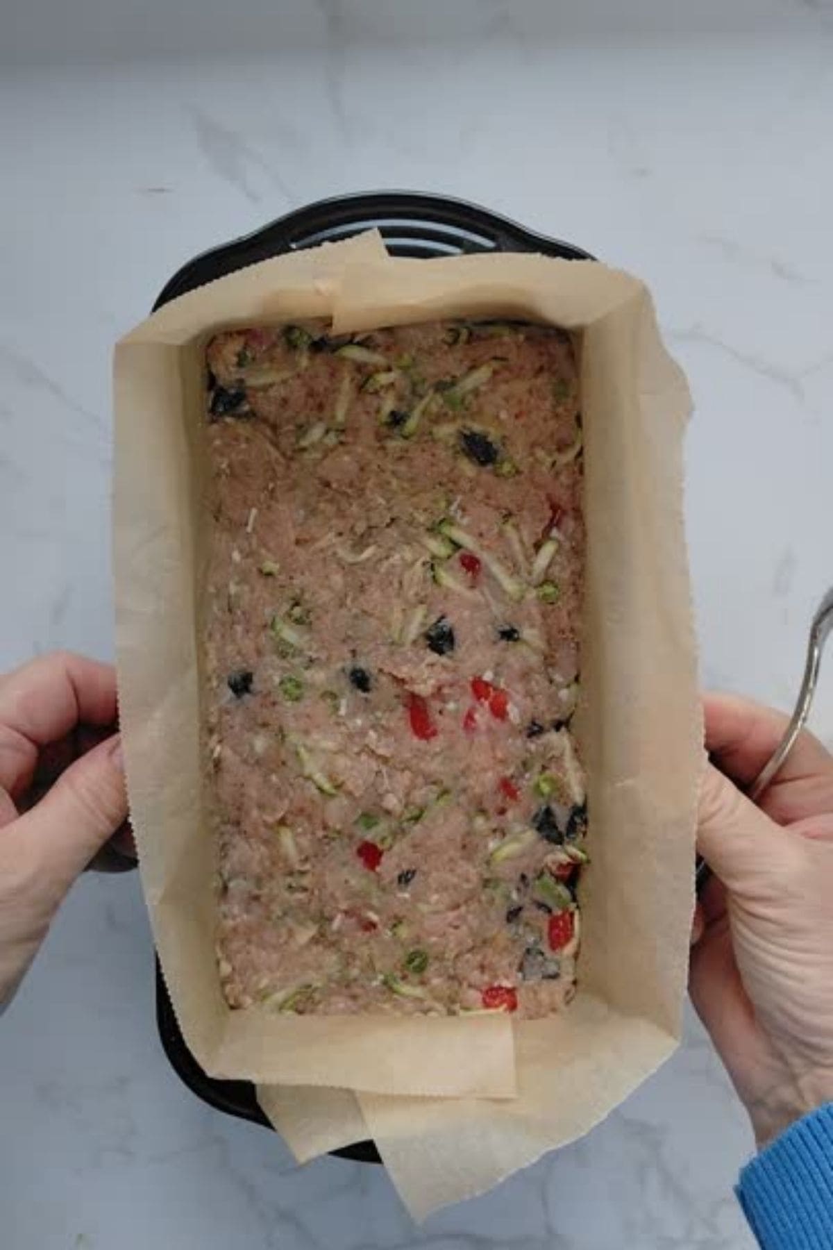 A pair of hands hold a loaf pan lined with parchment paper, filled with an uncooked chicken meatloaf for dogs, featuring chopped vegetables.
