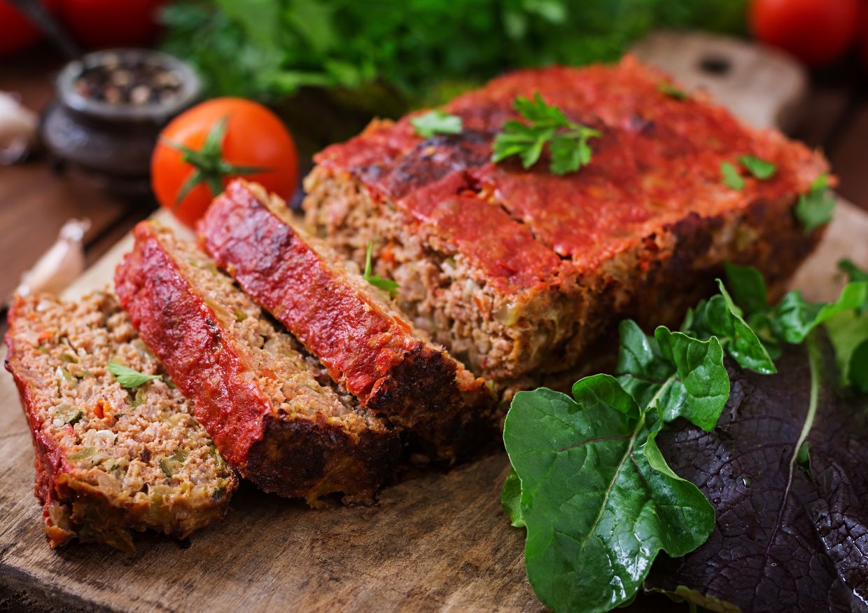 Sliced meatloaf with a tomato glaze on a wooden board, garnished with parsley and served with fresh leafy greens and a tomato.