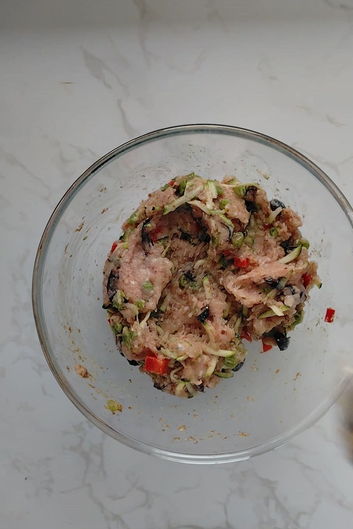 A glass bowl containing a mixture of ground meat, chopped vegetables, and seasonings sits on a white marble surfaceโperfect for preparing a wholesome chicken meatloaf for dogs.