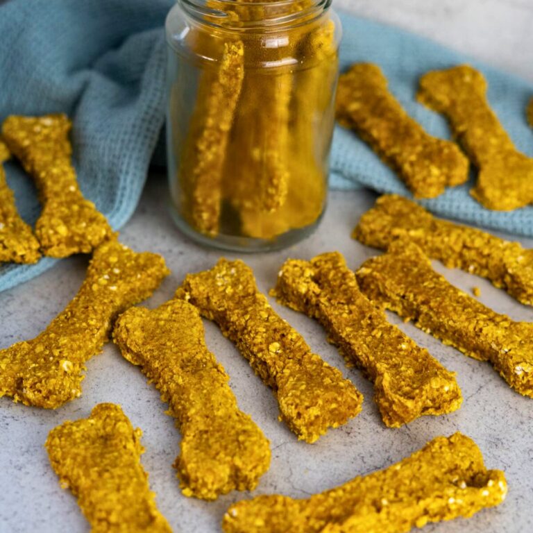 Bone-shaped homemade dog treats are arranged on a surface around a glass jar and a blue cloth.