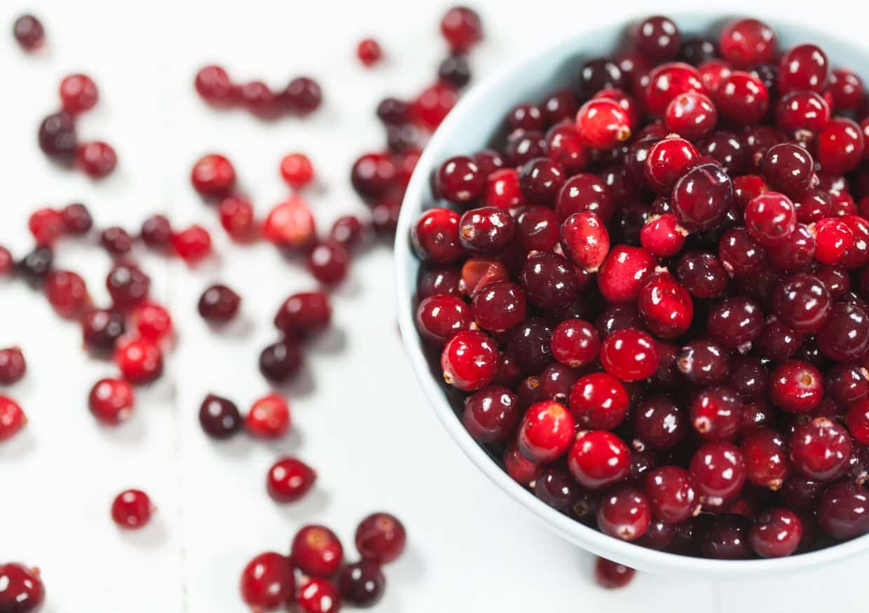 A bowl filled with fresh cranberries sits on a white surface, with some cranberries scattered around the bowl.