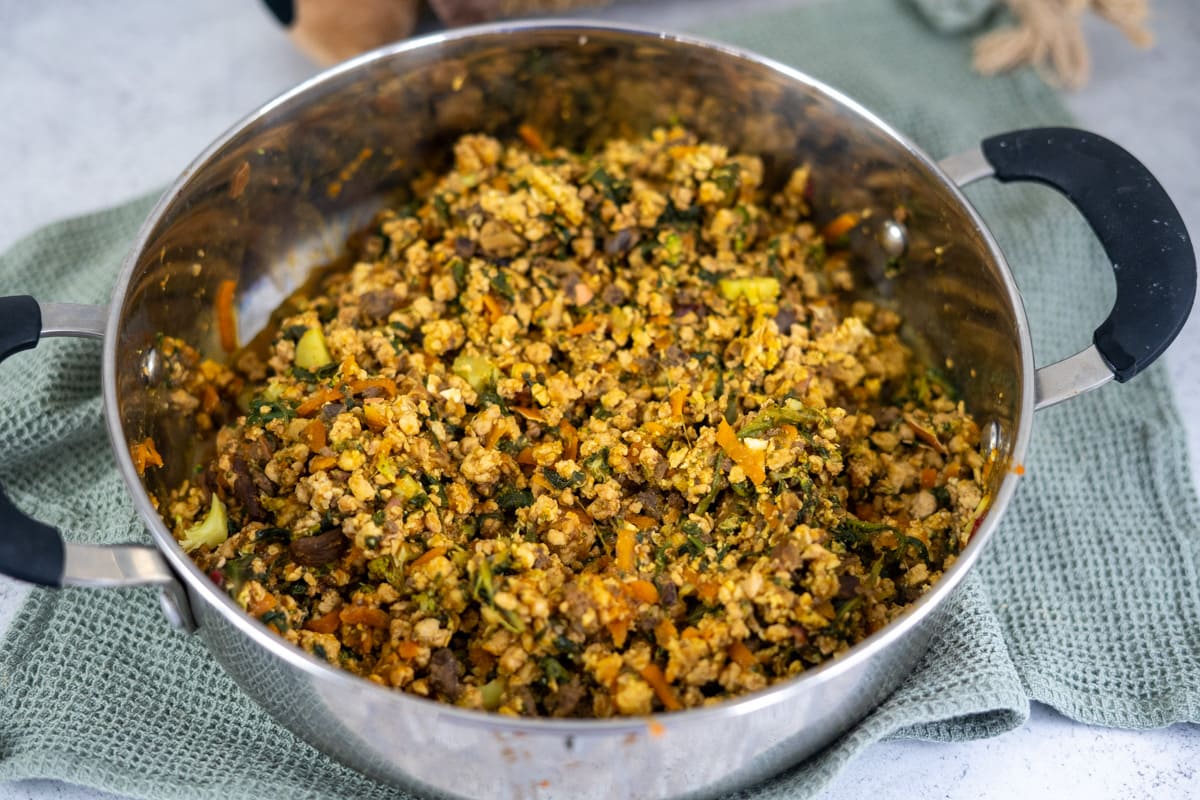 A stainless steel pot filled with a cooked mixture of vegetables, tofu, and possibly grains, resting on a green textured cloth.