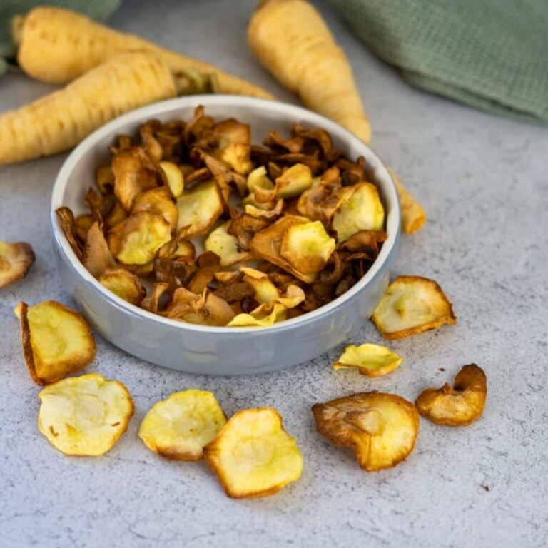 A bowl of parsnip chips with several chips scattered on a gray surface and whole parsnips in the background.