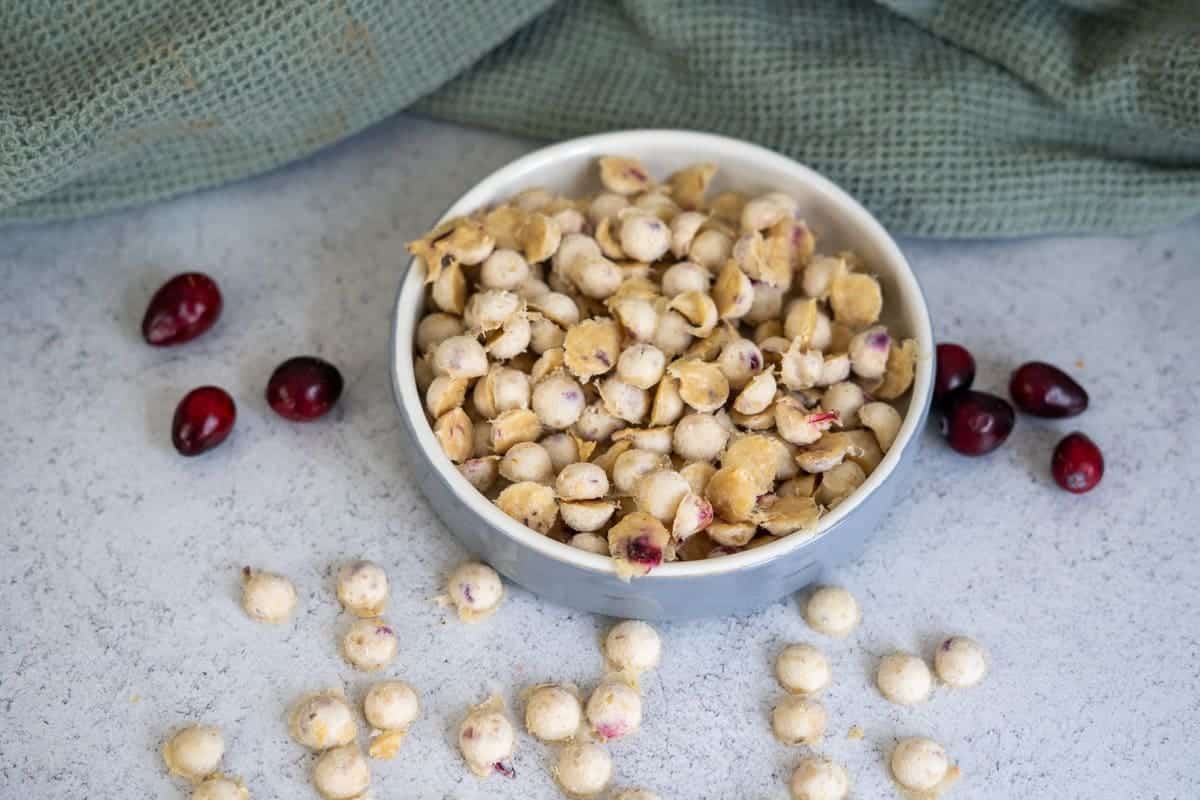 A white bowl filled with yogurt-covered cranberries sits on a light surface, with some cranberries and snacks scattered around and a green cloth in the background.