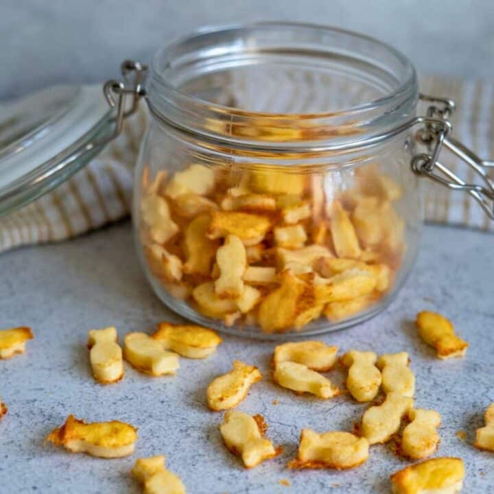 An open glass jar filled with fish-shaped cheese crackers sits on a gray surface with some crackers scattered outside the jar.