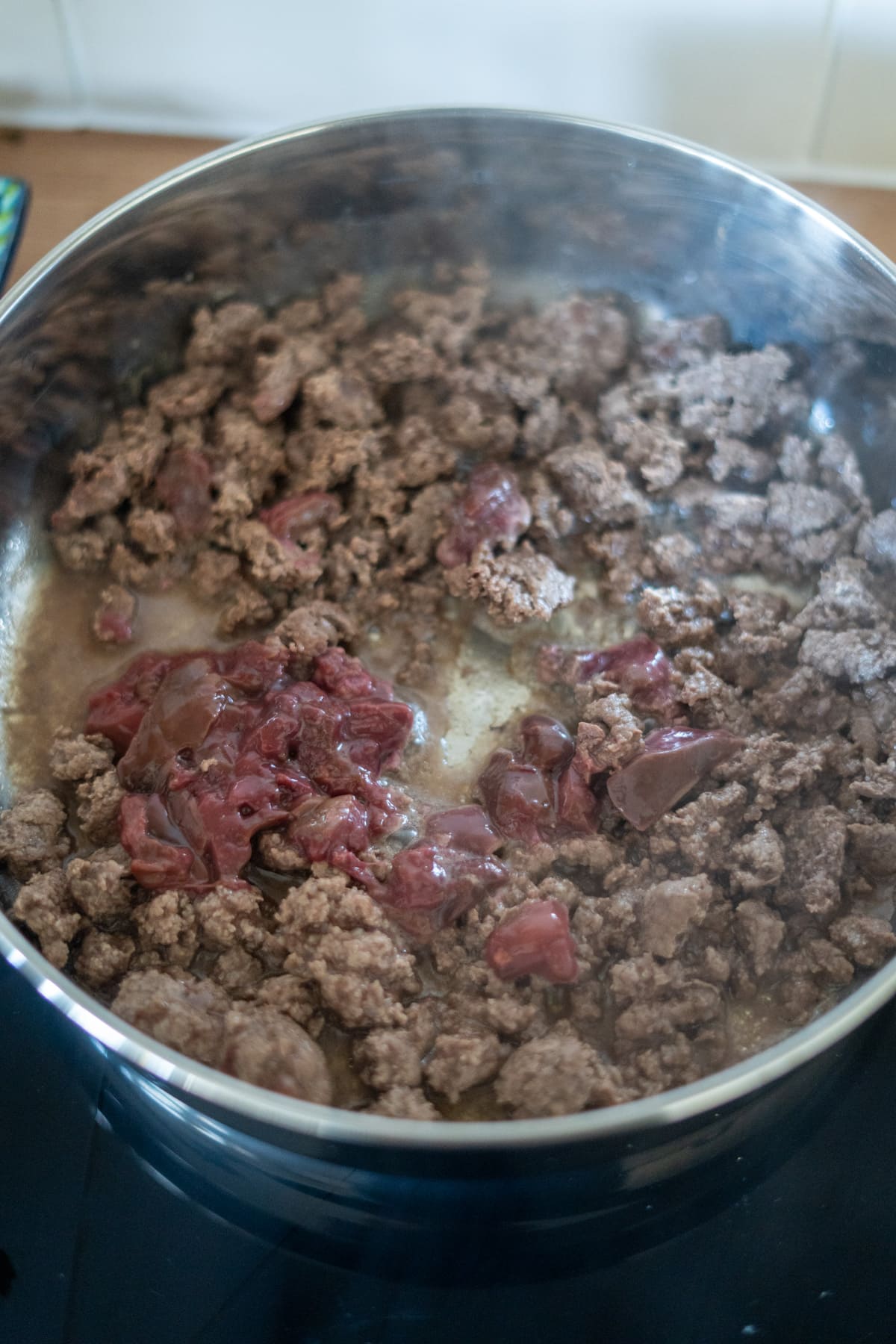 Minced beef and pieces of raw liver are being cooked together in a stainless steel saucepan on a hob as part of a nutritious venison dog food recipe.