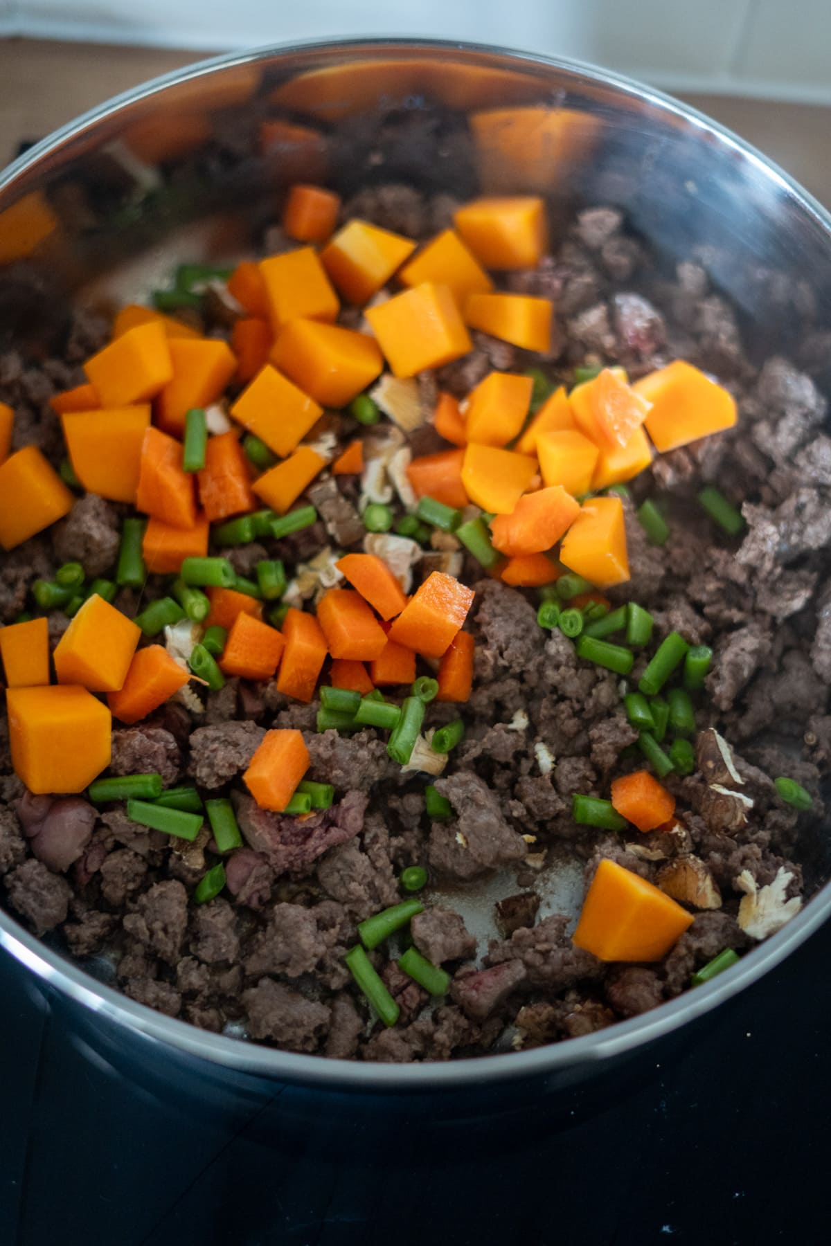 Minced beef, chopped orange pumpkin, and green beans are cooking together in a stainless steel saucepan on the hob, creating a savoury mix perfect as inspiration for a homemade venison dog food recipe.