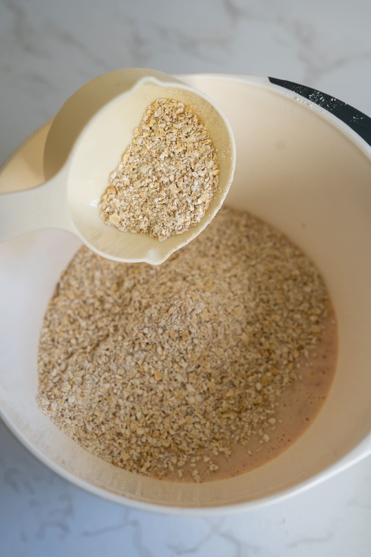 A white measuring jug holds rolled oats above a mixing bowl containing more oats and a liquid mixture, ready to be combined for delicious strawberry banana pupcakes on a light worktop.