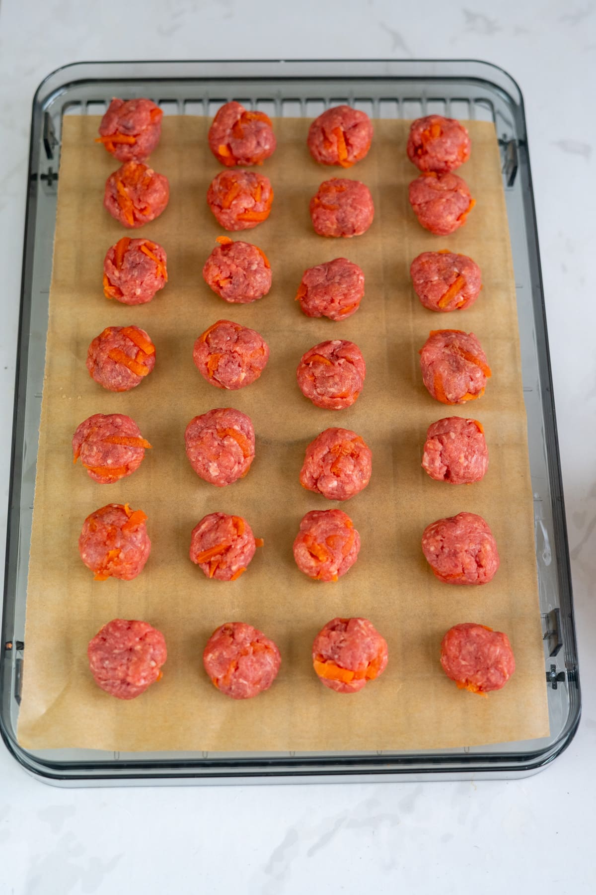 Rows of raw meatballs with visible orange pieces are arranged on parchment paper atop a wire rack, ready for cooking.