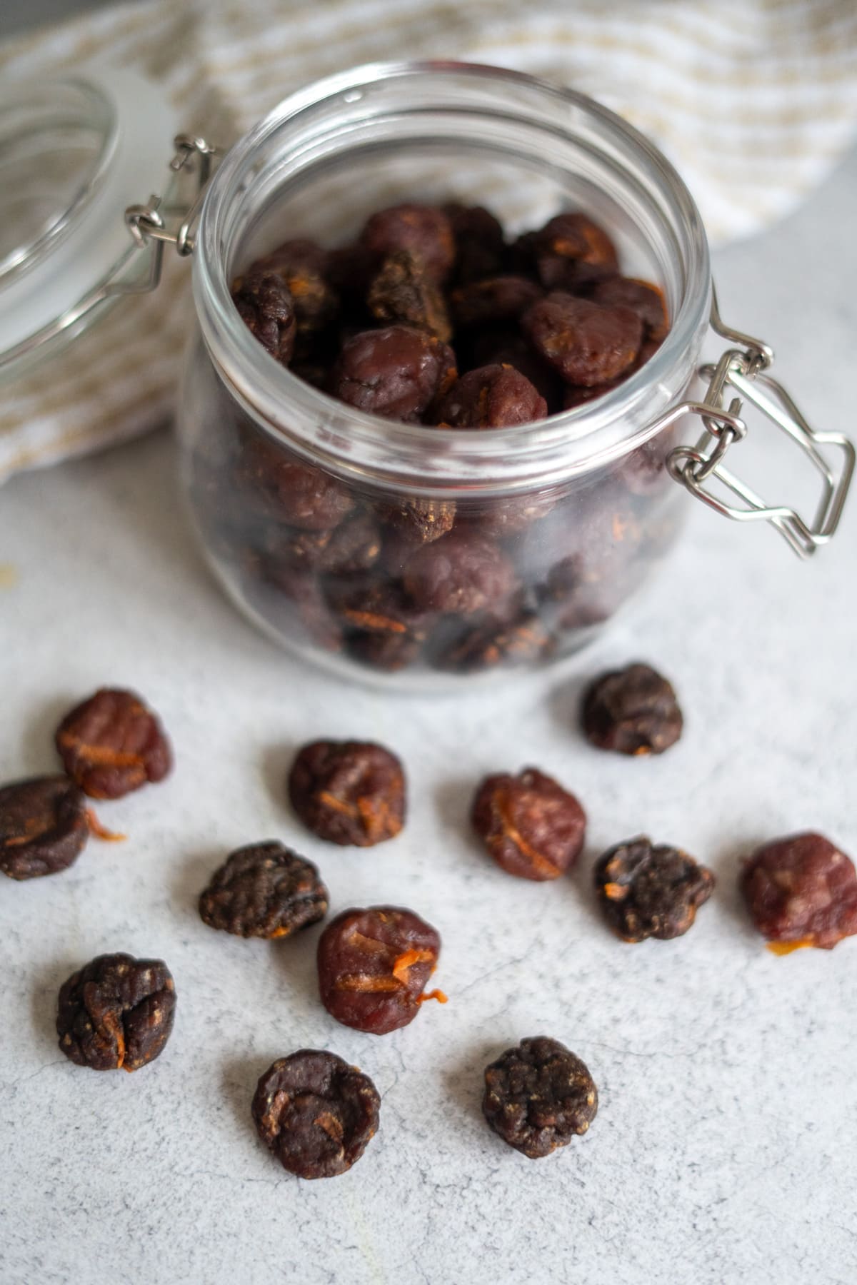 A glass jar filled with dried plums, with several dried plums scattered on a light-colored surface in front of the jar.