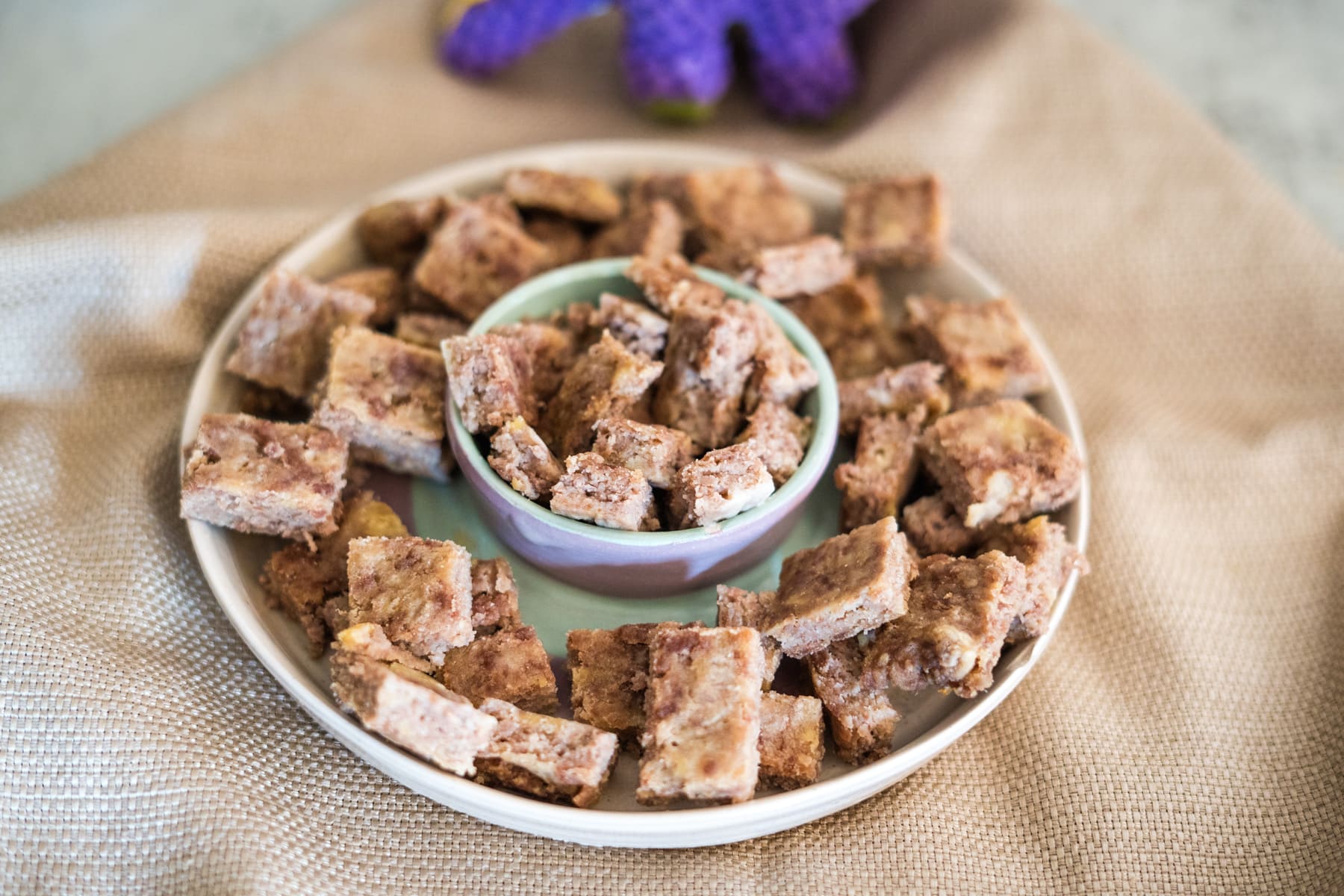 A plate with small square pieces of a brown, nutty snack, arranged around and inside a small bowl, placed on a light tan fabric surface.