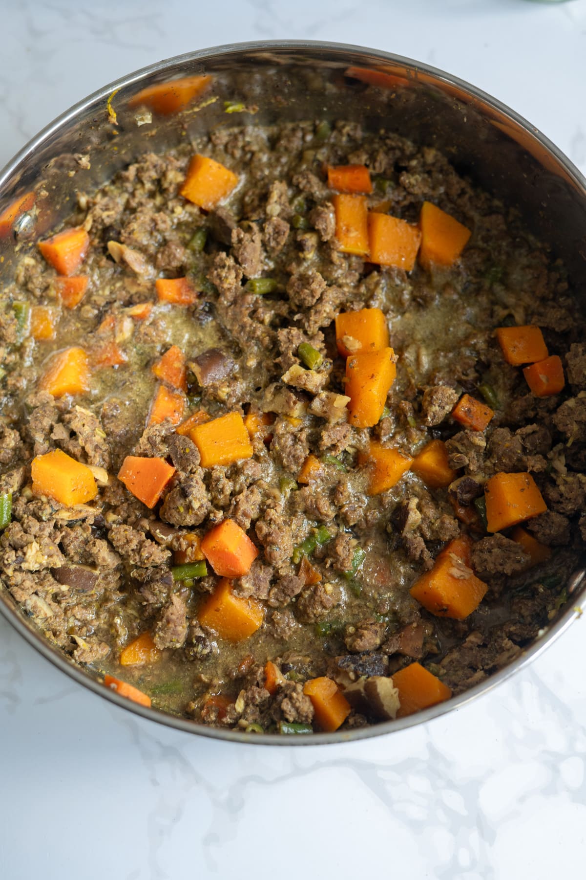 A pot filled with cooked minced venison, diced orange sweet potatoes, green vegetables, and a light broth—an appetising venison dog food recipe—sits on a white worktop.