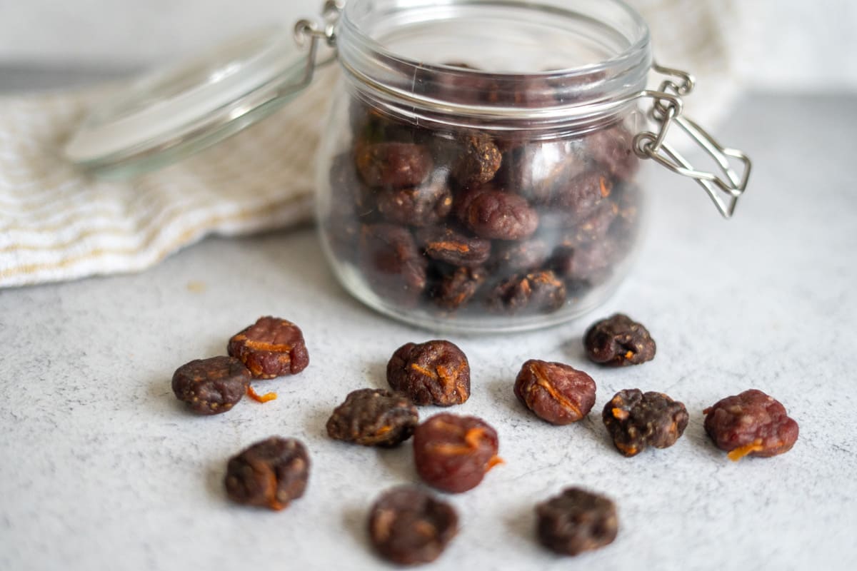 A glass jar filled with dried plums, with several dried plums scattered on a light gray surface in front of it.