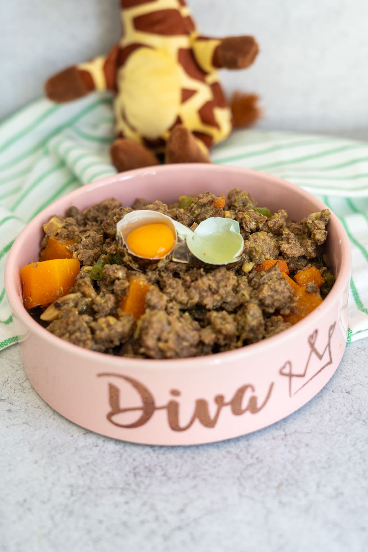 A pink pet bowl labelled "Diva" filled with a venison dog food recipe of minced meat, diced vegetables, and two egg yolks, with a stuffed giraffe toy and striped cloth in the background.