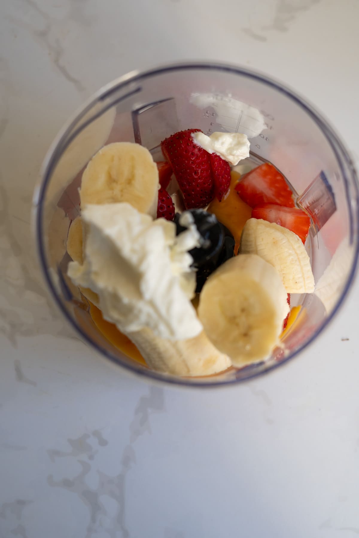 A blender jug containing sliced bananas, strawberries, dollops of yoghurt, and orange juice, ready to be blended into a delicious base for strawberry banana pupcakes.