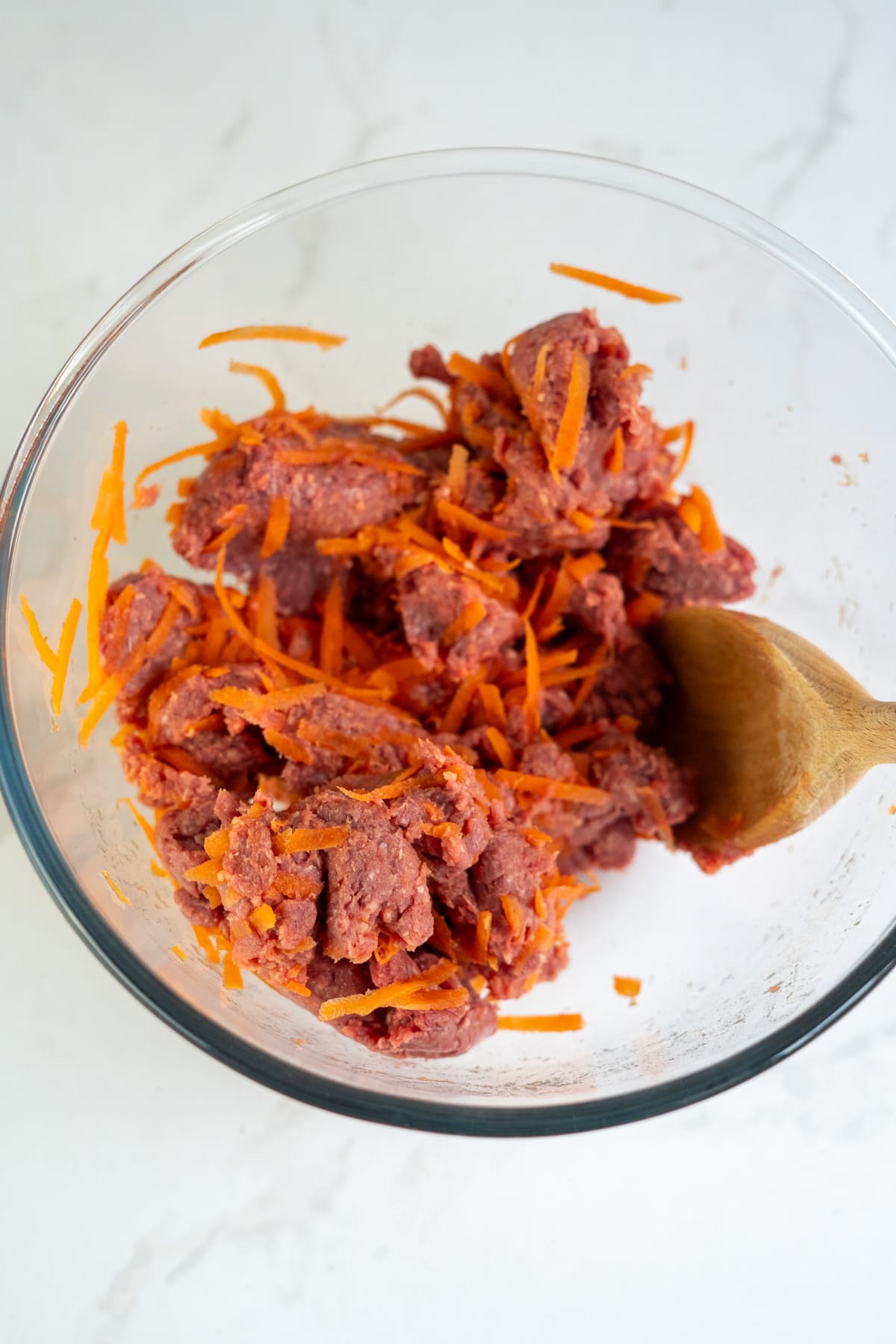 Ground beef and shredded carrots are mixed together in a clear glass bowl with a wooden spoon on a white surface.