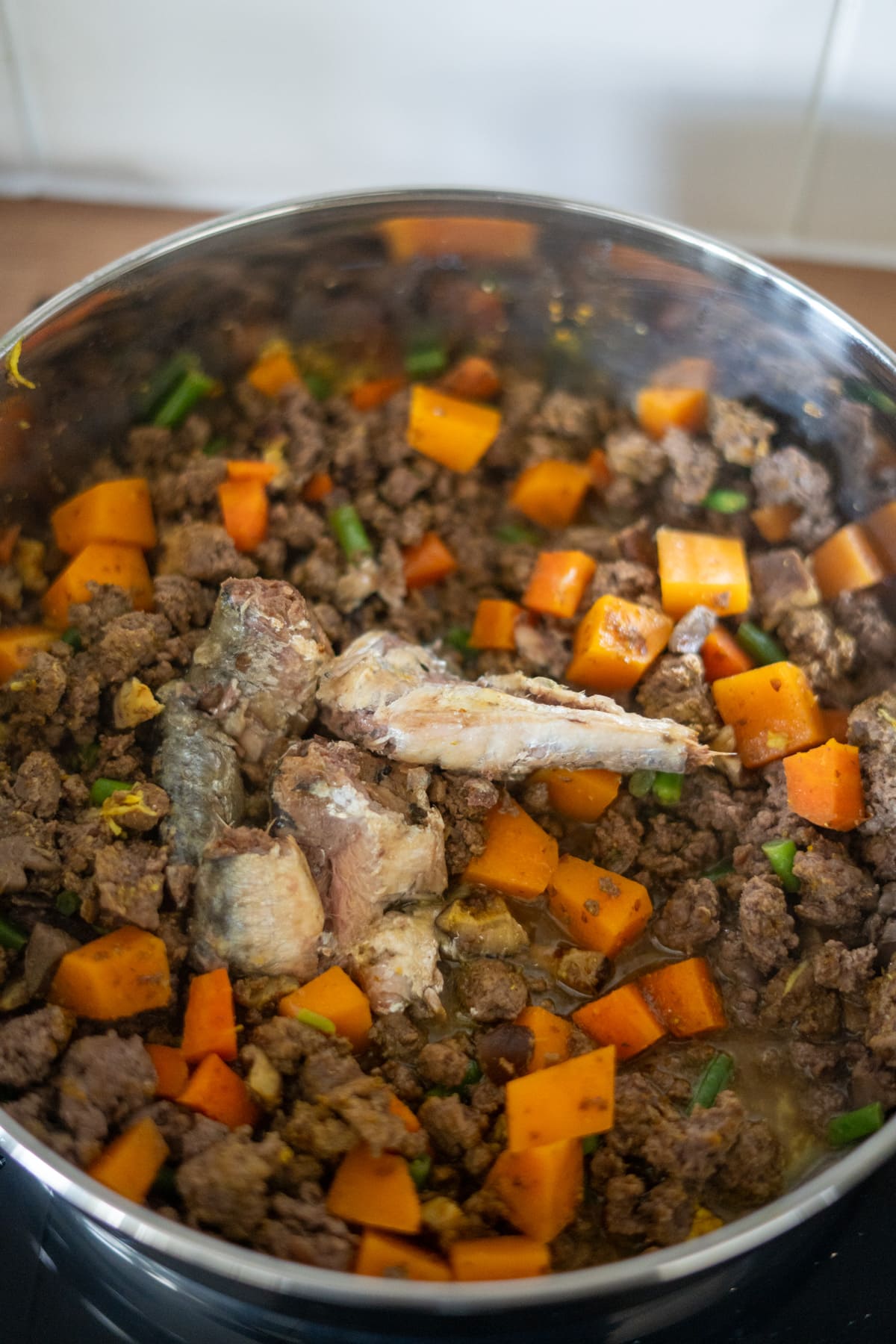 A pot of cooked minced meat, chopped sweet potatoes, green beans, and several pieces of fish—all mixed together on a hob for a hearty venison dog food recipe.