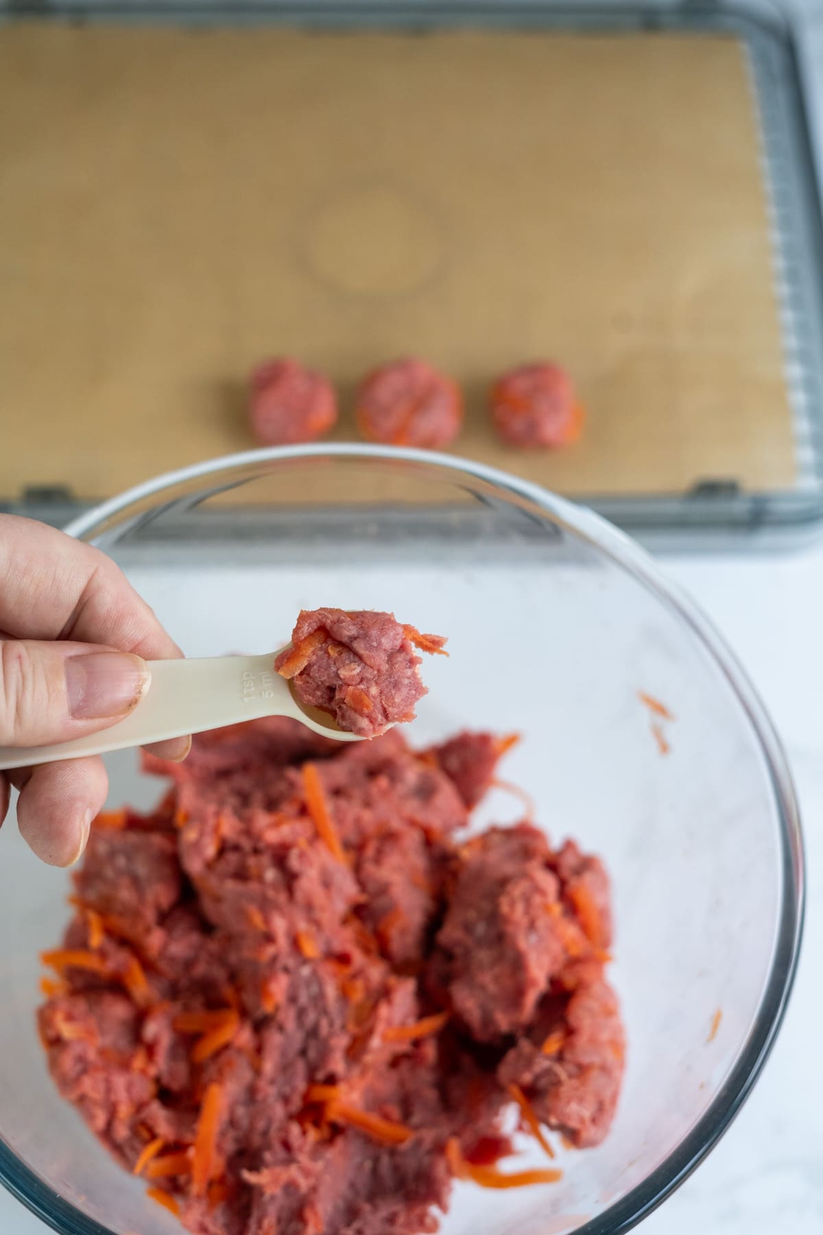 A hand holds a spoonful of ground meat and shredded carrots mixture over a bowl, with small meatballs arranged on a tray in the background.