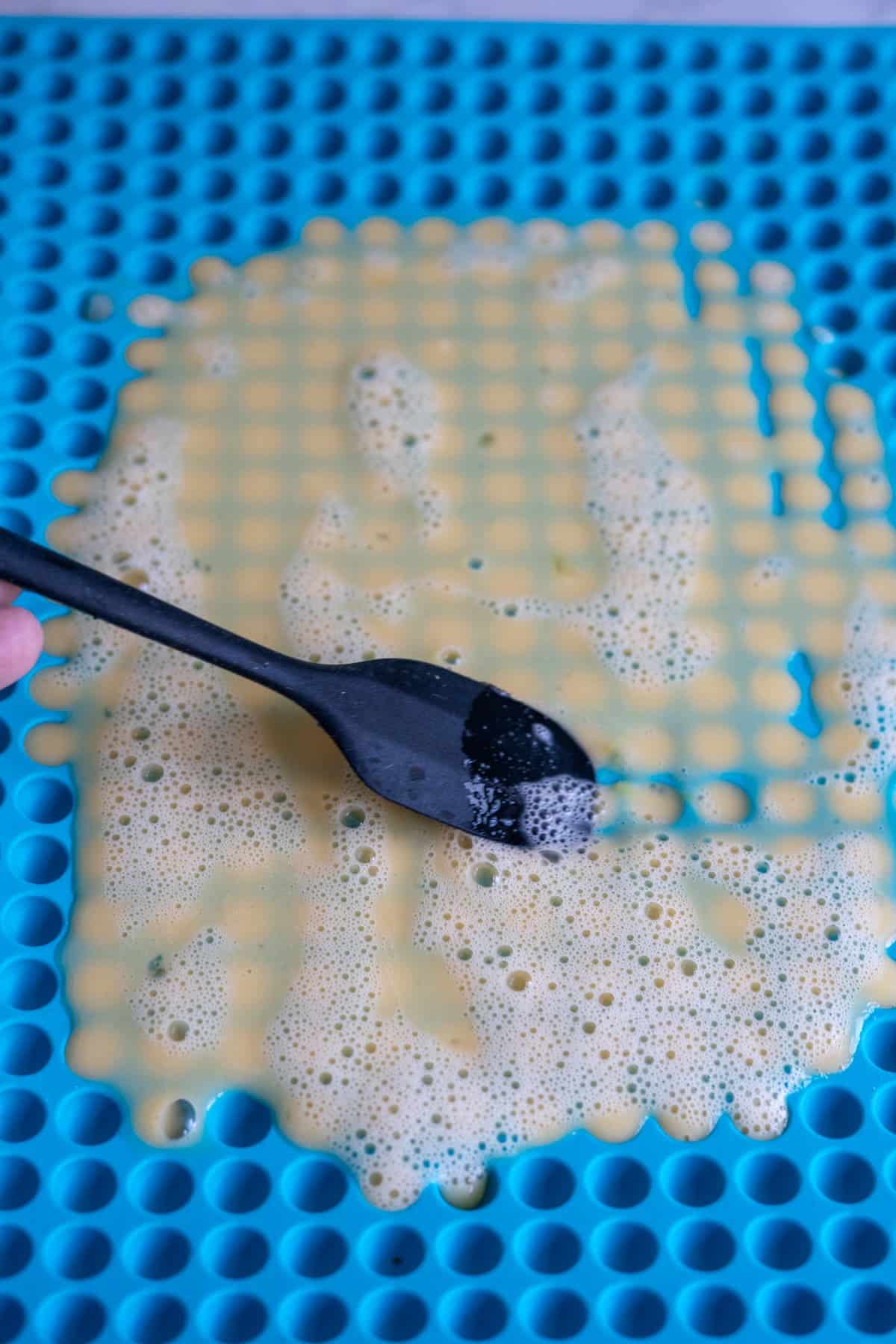 A black spoon spreads a liquid mixture for dog egg treats over a blue silicone baking mat with raised circular molds.