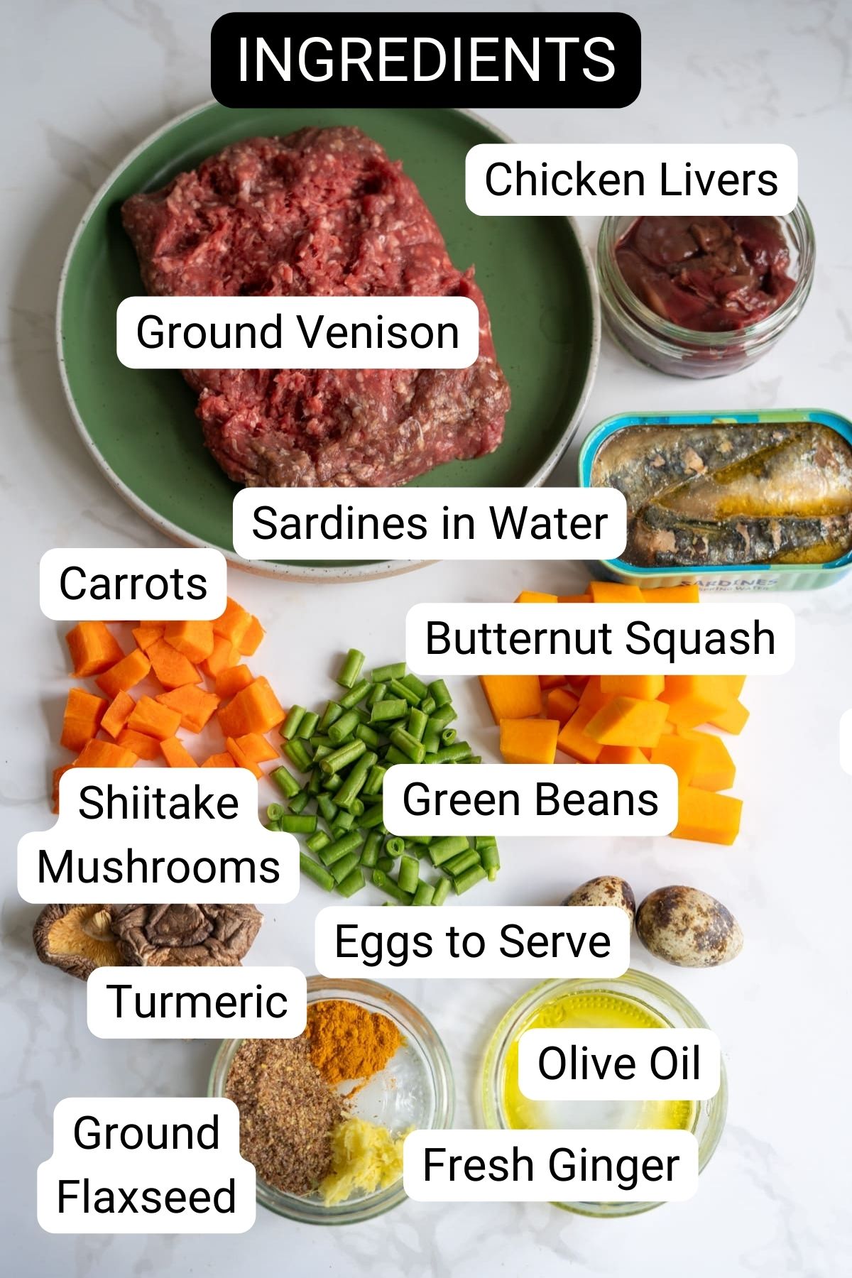 A variety of labelled ingredients on a white surface, including minced venison, ideal for a nutritious venison dog food recipe, plus chicken livers, sardines, carrots, butternut squash, mushrooms, green beans, turmeric, eggs, olive oil, linseed, and ginger.