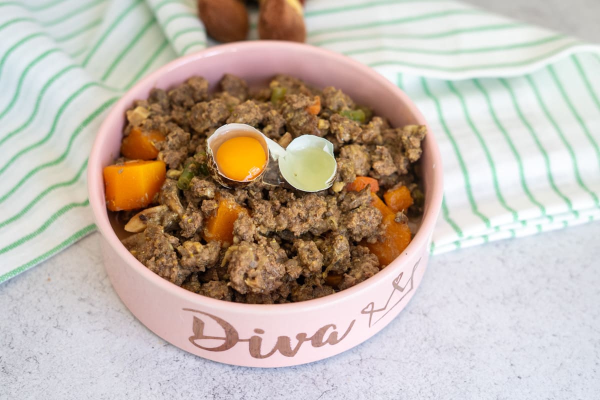A pink bowl labelled “Diva” filled with venison dog food recipe—minced meat, vegetables, and two cracked quail eggs—sits on a worktop beside a green-striped cloth.