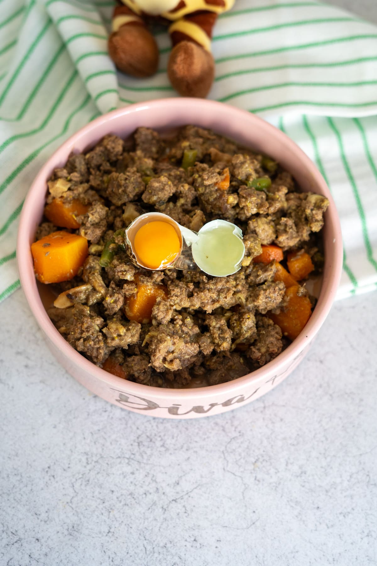 A bowl of homemade venison dog food recipe with minced meat, vegetables, a raw egg yolk, and green liquid on top, set on a striped cloth with a soft toy in the background.