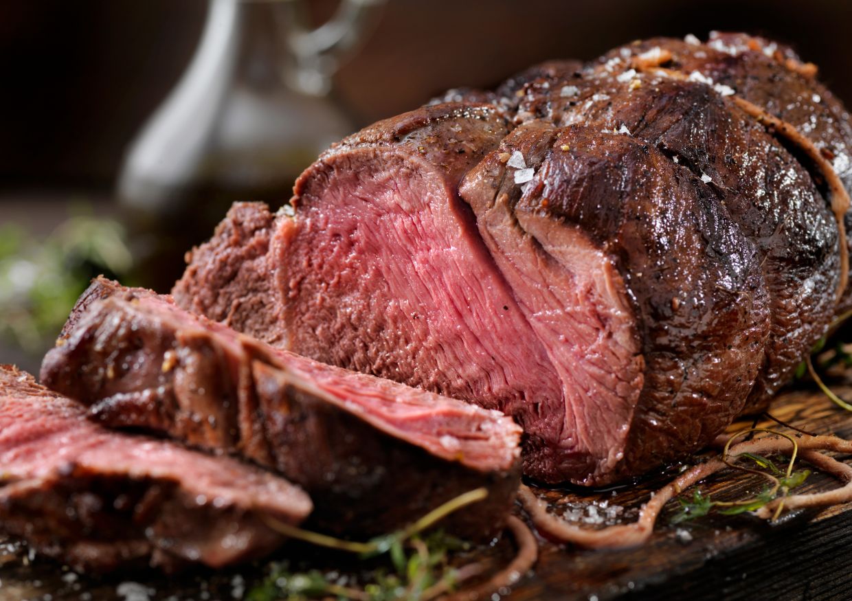 A close-up of a sliced medium-rare roast beef on a wooden board, with visible seasoning and fresh herbs.