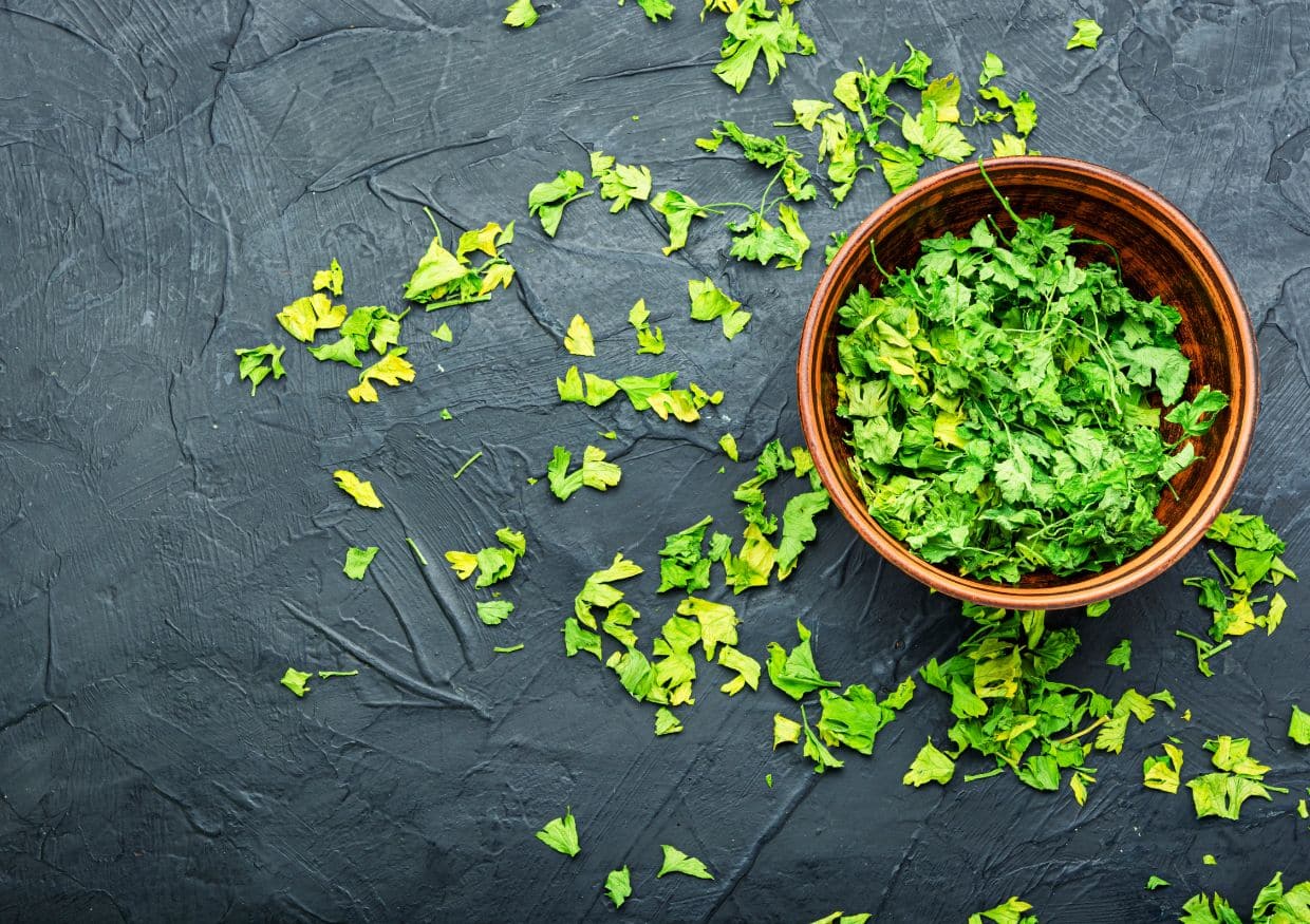 A wooden bowl filled with chopped fresh parsley sits on a dark textured surface with scattered parsley leaves around it.