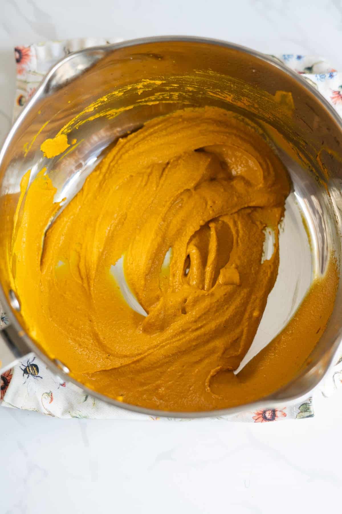 A stainless steel mixing bowl containing smooth, orange pumpkin purée sits on a patterned cloth on a white surface.