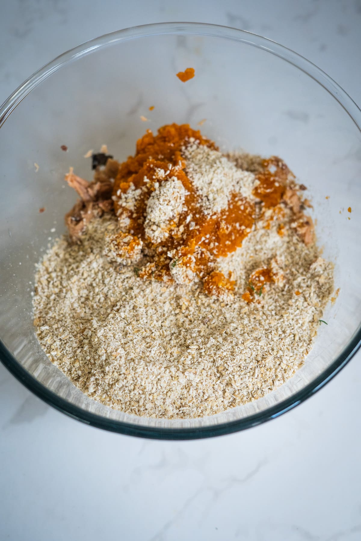A glass bowl containing a mixture of orange purée, breadcrumbs, and shredded ingredients—perfect for making salmon meatballs for dogs—sits on a white worktop.