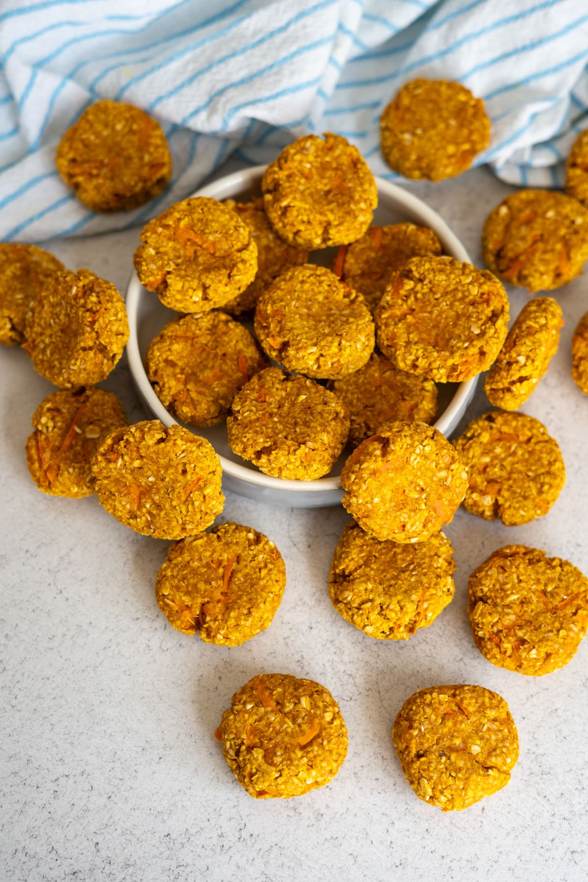A bowl of round, golden pumpkin carrot dog biscuits with several biscuits scattered on a light worktop, next to a blue and white striped tea towel.