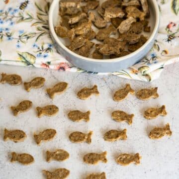 Small fish-shaped biscuits are arranged on a light surface, with more biscuits in a round dish placed on a floral serviette.
