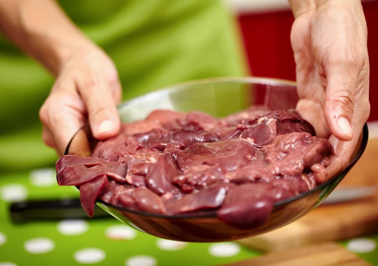 A person holds a glass bowl filled with raw chicken liver for dogs over a green spotty tablecloth.