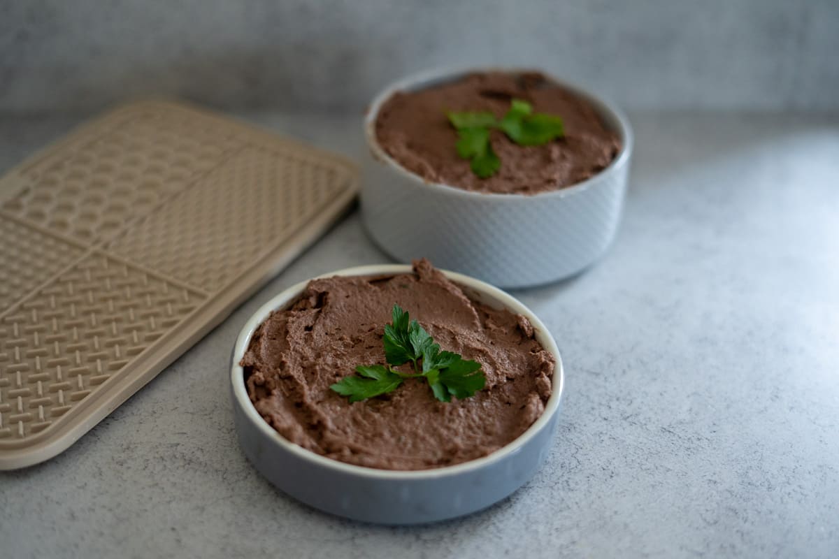 Two small bowls filled with a brown chicken liver pâté for dogs, each garnished with fresh parsley, placed on a light grey surface next to a beige textured tray.