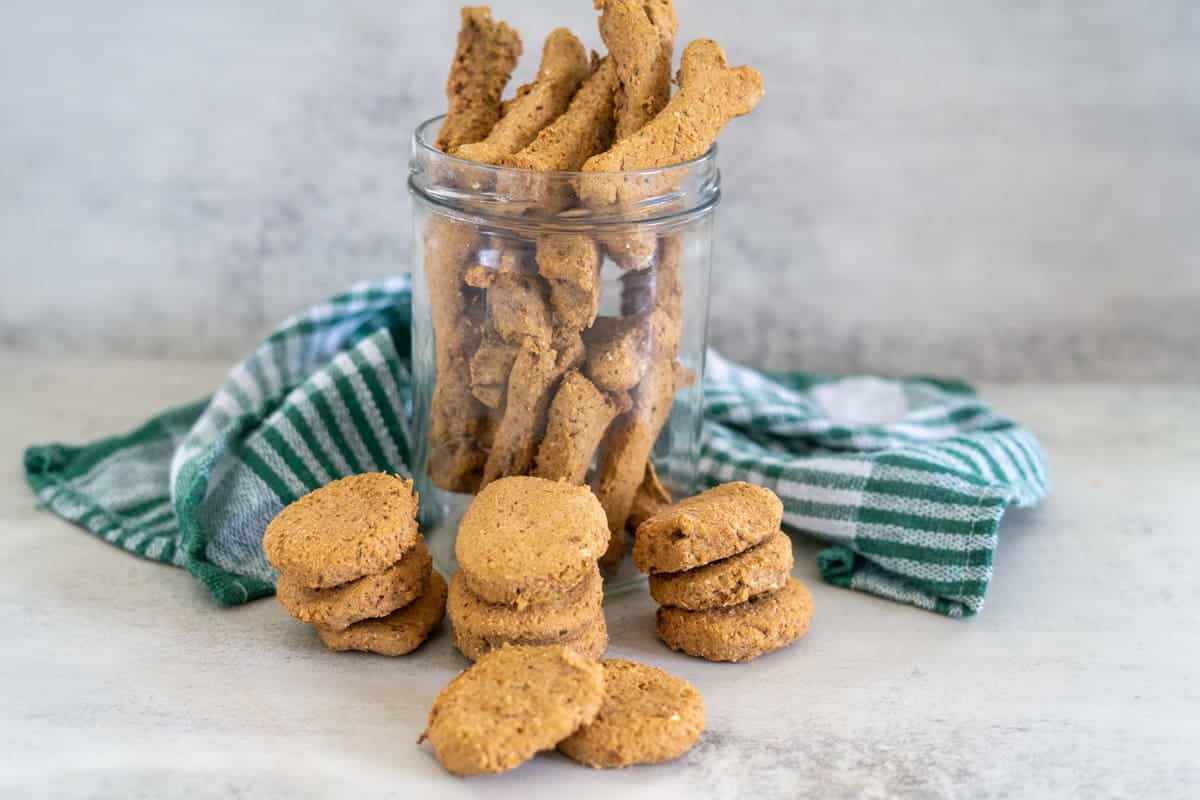 A glass jar filled with homemade dog biscuits made with chicken liver for dogs sits on a counter, with more biscuits stacked and scattered in front, and a green striped cloth in the background.