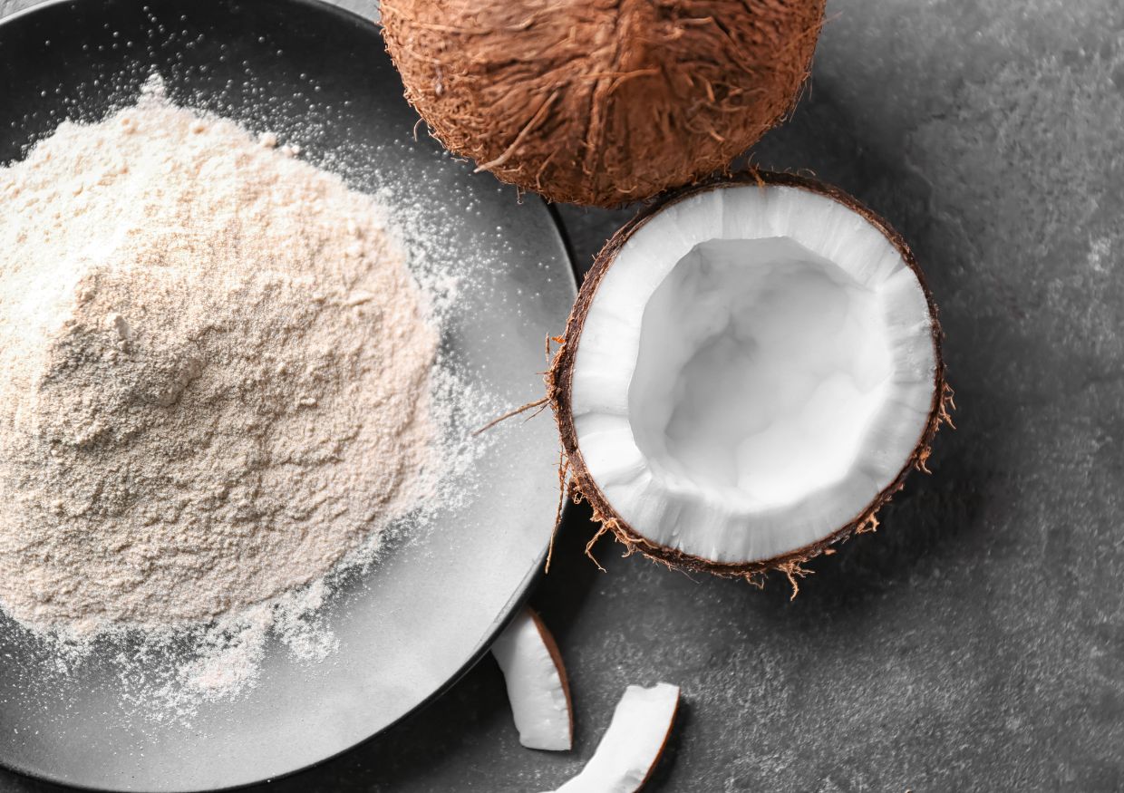 A plate of coconut flour next to a whole coconut, a halved coconut with white flesh, and coconut pieces on a dark surface, perfect for those wondering: can dogs eat coconut flour?.