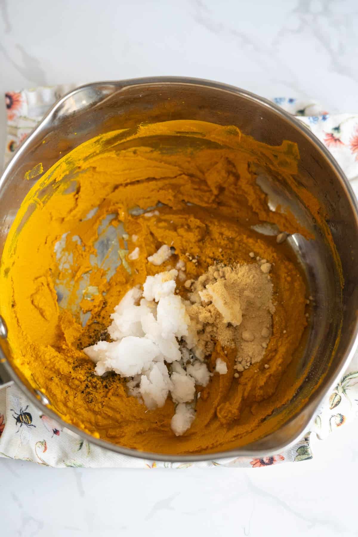 Stainless steel bowl with a yellow mixture, coconut oil, ground spices, and pepper on a patterned cloth, on a white worktop.