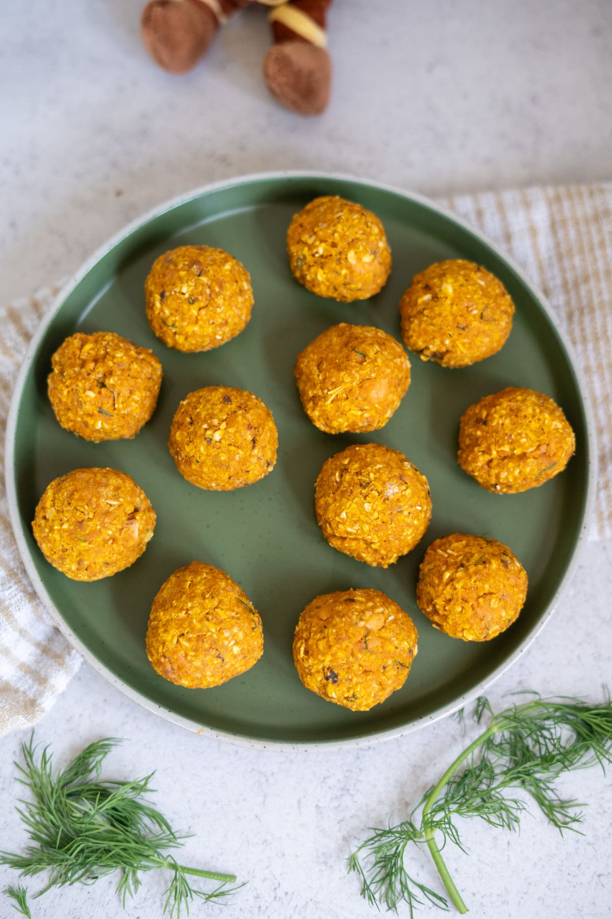 A green plate with twelve round, orange-coloured salmon meatballs for dogs sits on a light surface, with a striped cloth and sprigs of dill nearby.